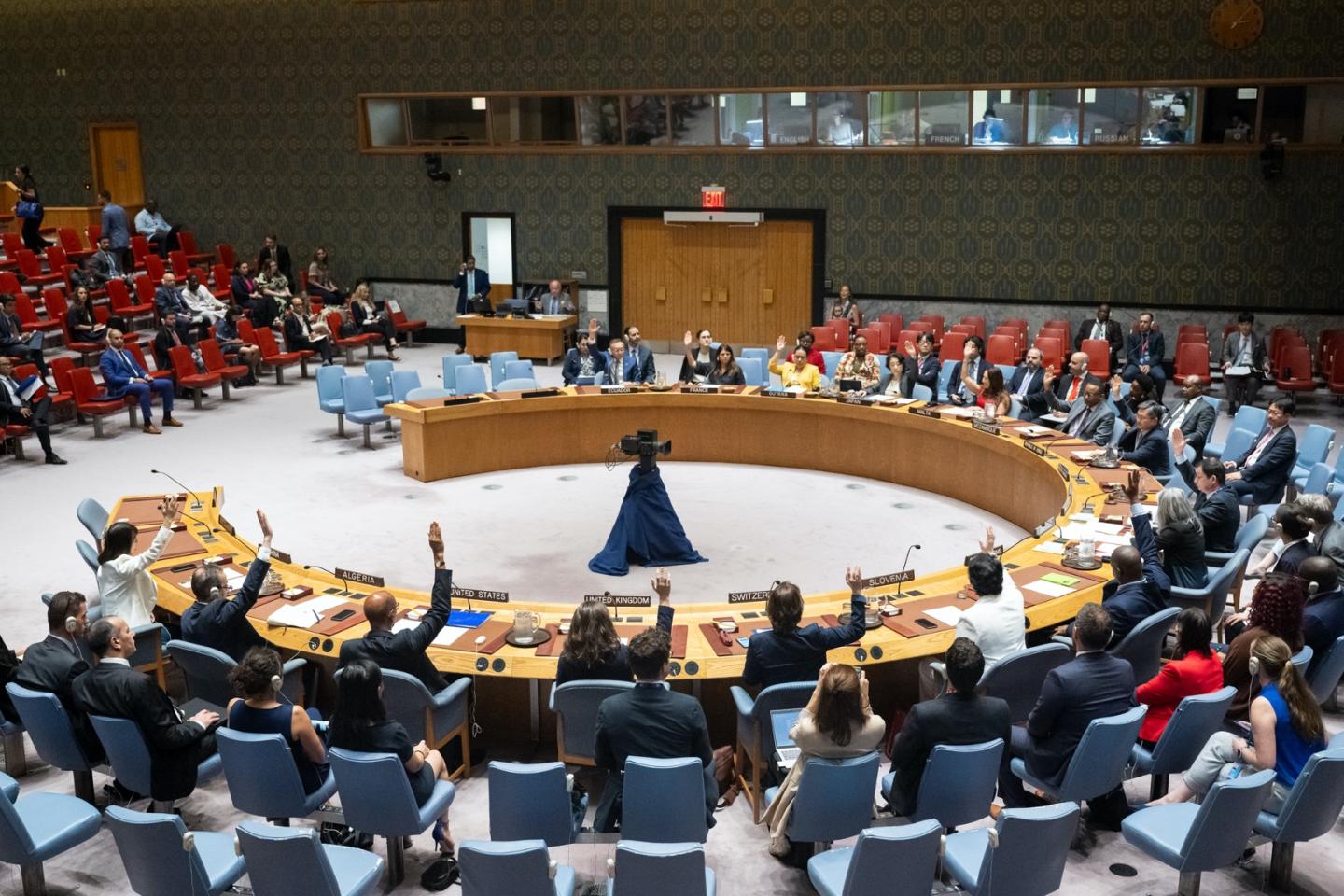 Wide view of meeting in session with several attendees raising hands.